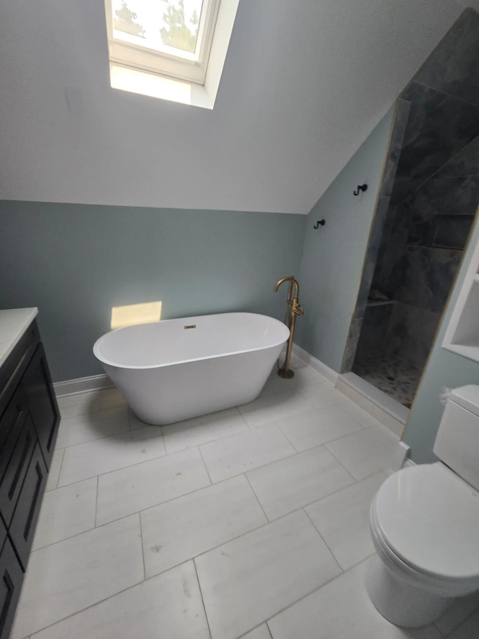 Freestanding white soaking tub with bronze filler, white subway tile floor, skylight, and updated bathroom plumbing