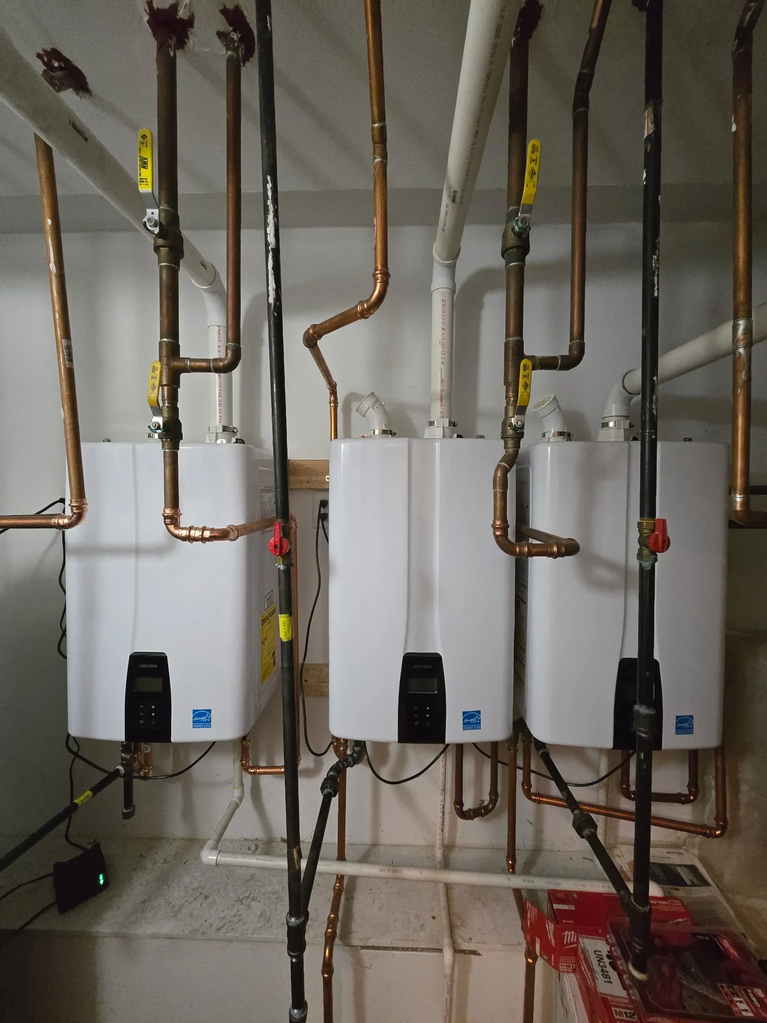 Three white tankless water heaters with copper piping, yellow valve markers, and white vent lines installed on wall