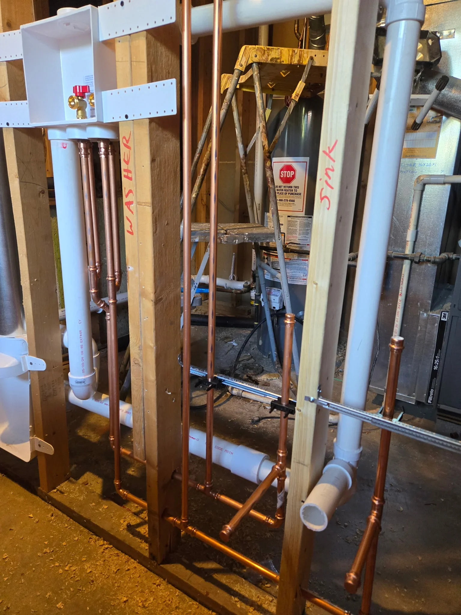 Copper plumbing lines with soldered joints and white PVC drain pipes installed in framed wall cavity
