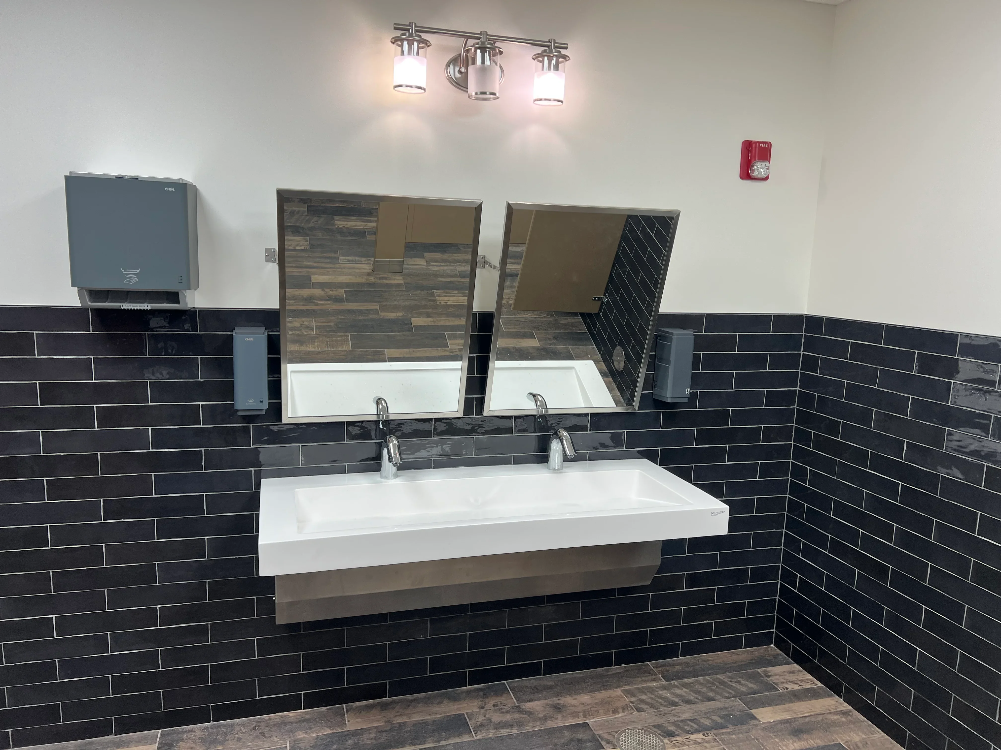 Dual-sink commercial vanity with white countertop, chrome faucets, and black subway tile walls