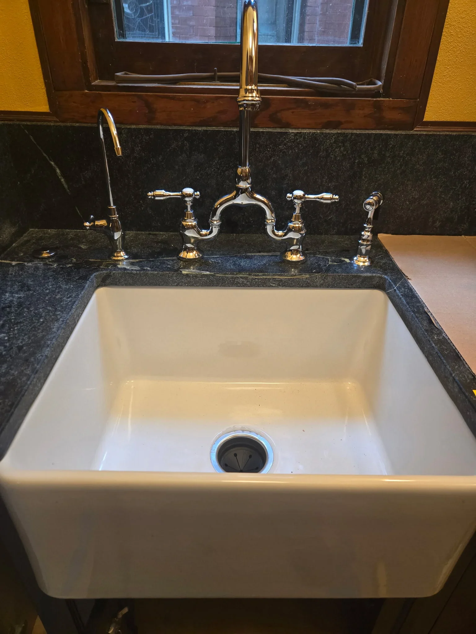 White ceramic farmhouse sink with polished brass bridge faucet and dual handles on dark granite countertop