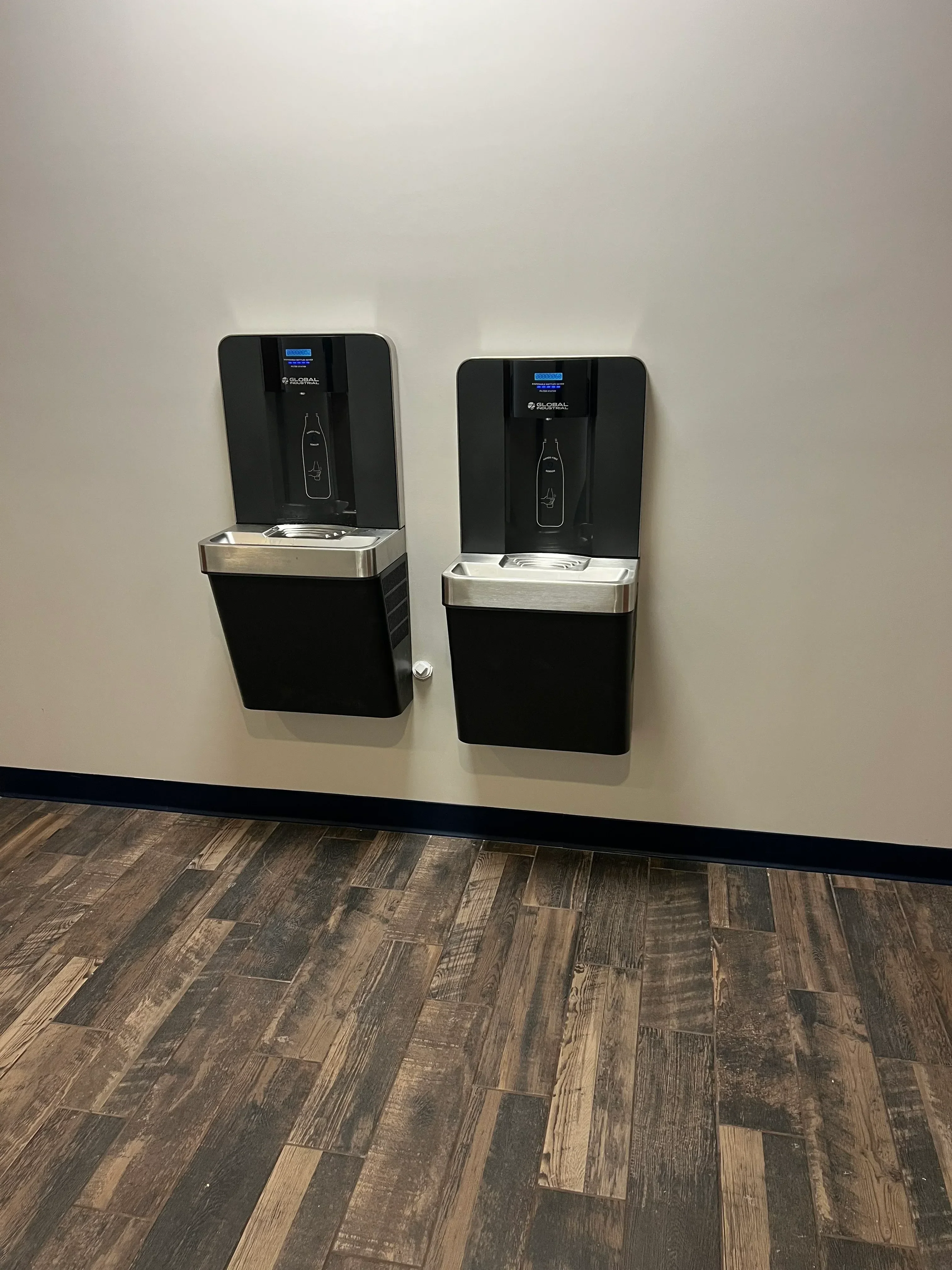 Two black tankless water heaters with stainless steel trim mounted on white wall beside wood-look tile